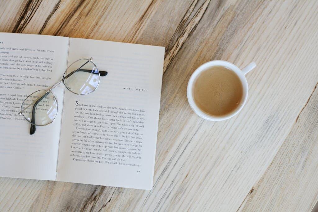 A calming flat lay image of a book, eyeglasses, and coffee on a wooden table.
