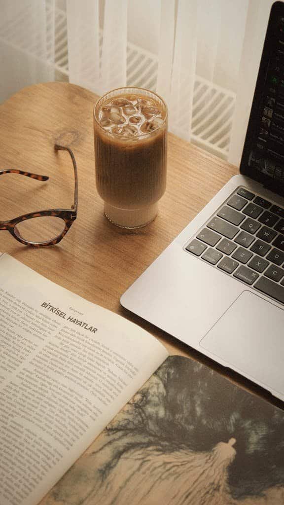 A serene workspace with iced coffee, open book, and laptop, perfect for work or study.